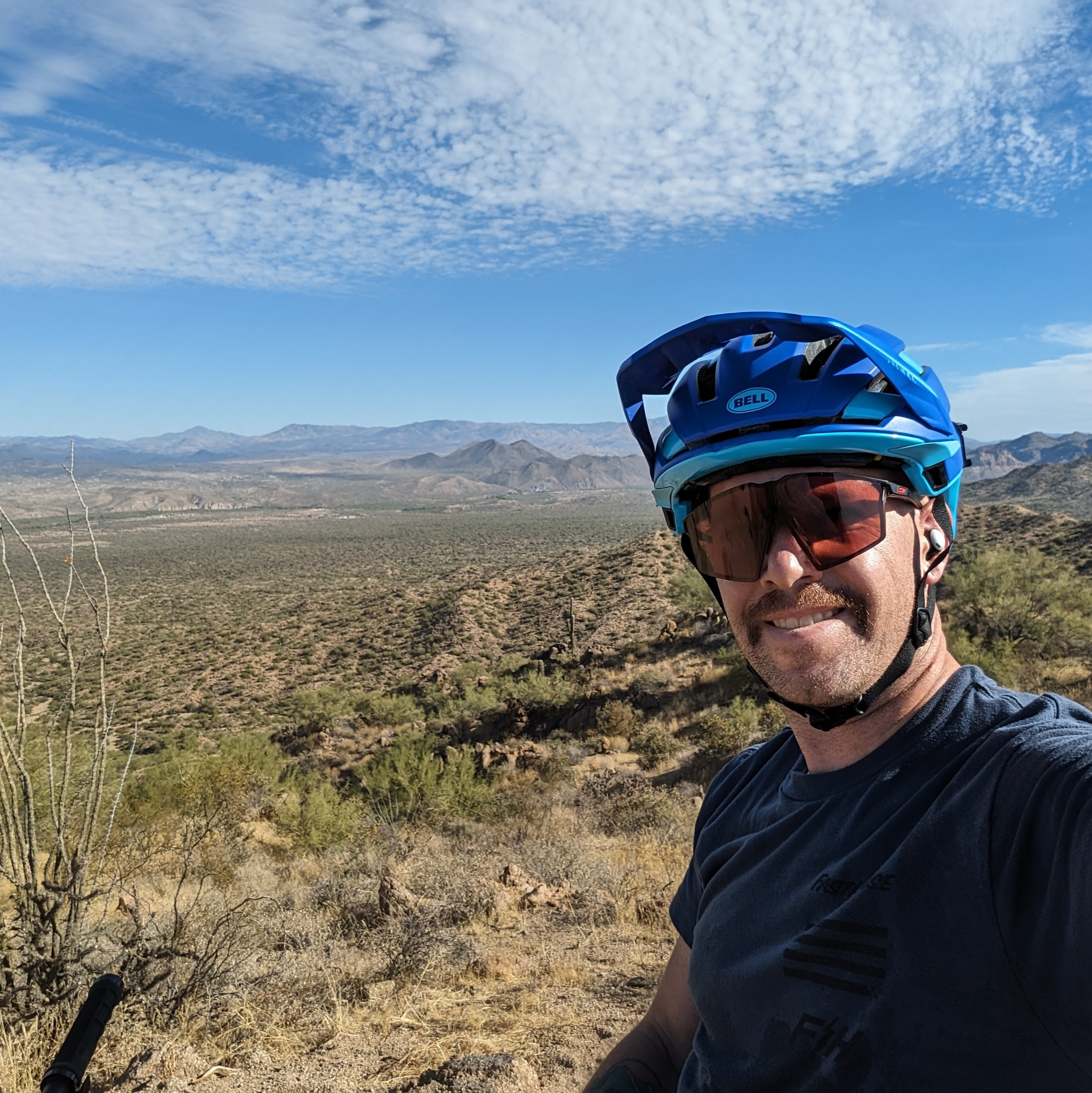 A selfie picture of Josh Lau with a mountain biking helmet and sunglasses on the top of a trail with blurred mountains in the background